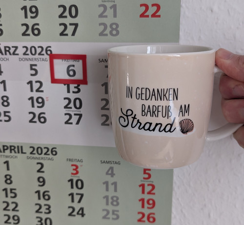 Meine rechte Hand hält einen Porzellan-Kaffeebecher. Seine Farbe ist beige. In schwarzer Schrift steht auf ihm: In Gedanken barfuß am Strand, gefolgt von einer Zeichnung einer bräunlichen Meermuschel. Im Hintergrund hängt ein 3-Monats-Wandkalender an der Wand mit dem rot umrandeten heutigen Tag. 