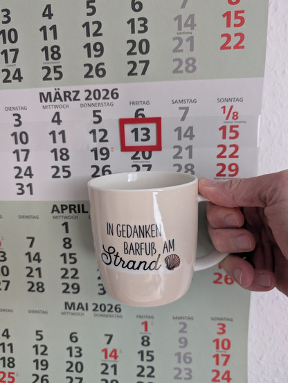 Meine rechte Hand hält einen Porzellan-Kaffeebecher. Seine Farbe ist beige. In schwarzer Schrift steht auf ihm: In Gedanken barfuß am Strand, gefolgt von einer Zeichnung einer bräunlichen Meermuschel. Im Hintergrund hängt ein 3-Monats-Wandkalender an der Wand mit dem rot umrandeten heutigen Tag. 