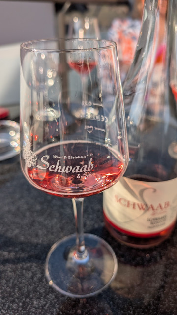In einem Weinglas mit der Aufschrift "Wein und Gästehaus Schwaab und Sohn" ist rötlicher Rosecco. Rechts daneben steht die Flasche mit dem Etikett mit derselben Aufschrift, tiefenunscharf. 