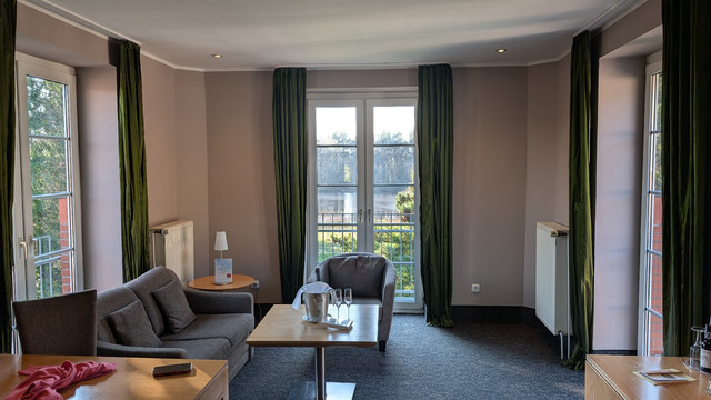 Eine Sofa-Sessel-Tisch-Sitzecke, die an drei Seiten von bodentiefen Fenstern umgeben ist. Groß. Viel Tageslicht. Aussicht ins Grüne.