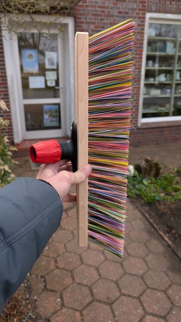 Meine linke Hand hält einen Besen ohne Stiel. Seine Borsten sind kunterbunt. Im Hintergrund ist die kleine Eingangstür zum Geschäft in Ihlowerfehn.