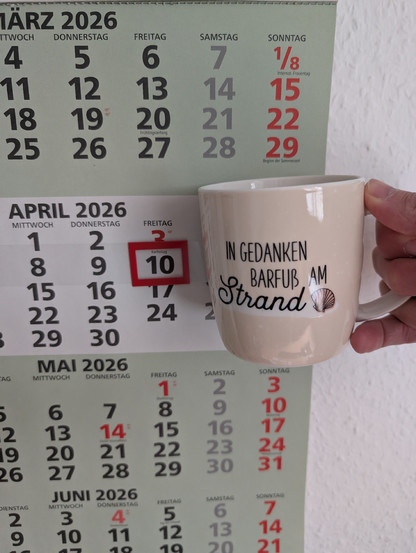Meine rechte Hand hält einen Porzellan-Kaffeebecher. Seine Farbe ist beige. In schwarzer Schrift steht auf ihm: In Gedanken barfuß am Strand, gefolgt von einer Zeichnung einer bräunlichen Meermuschel. Im Hintergrund hängt ein 3-Monats-Wandkalender an der Wand mit dem rot umrandeten heutigen Tag. 