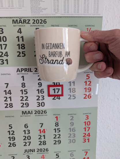 Meine rechte Hand hält einen Porzellan-Kaffeebecher. Seine Farbe ist beige. In schwarzer Schrift steht auf ihm: In Gedanken barfuß am Strand, gefolgt von einer Zeichnung einer bräunlichen Meermuschel. Im Hintergrund hängt ein 3-Monats-Wandkalender an der Wand mit dem rot umrandeten heutigen Tag. 