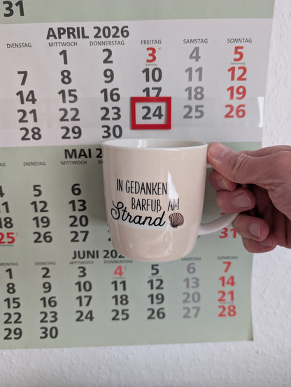 Meine rechte Hand hält einen Porzellan-Kaffeebecher. Seine Farbe ist beige. In schwarzer Schrift steht auf ihm: In Gedanken barfuß am Strand, gefolgt von einer Zeichnung einer bräunlichen Meermuschel. Im Hintergrund hängt ein 3-Monats-Wandkalender an der Wand mit dem rot umrandeten heutigen Tag. 