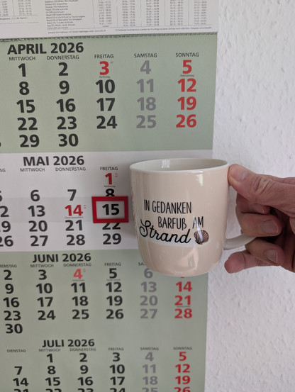 Meine rechte Hand hält einen Porzellan-Kaffeebecher. Seine Farbe ist beige. In schwarzer Schrift steht auf ihm: In Gedanken barfuß am Strand, gefolgt von einer Zeichnung einer bräunlichen Meermuschel. Im Hintergrund hängt ein 3-Monats-Wandkalender an der Wand mit dem rot umrandeten heutigen Tag. 