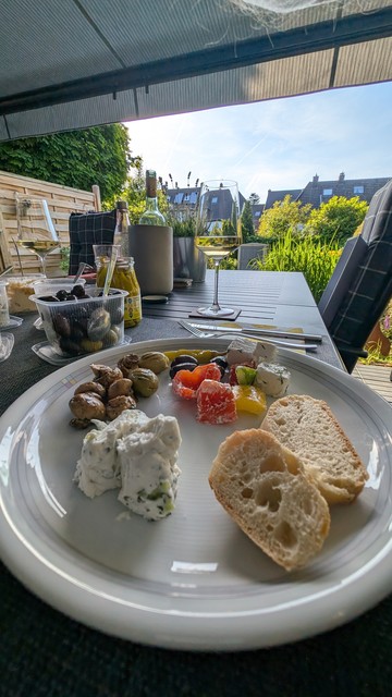 Im Vordergrund ein weißer Teller mit Antipasti und Brot. Dahinter ein Glas mit Weißwein. Im Hintergrund Garten und Nachbargebäude.