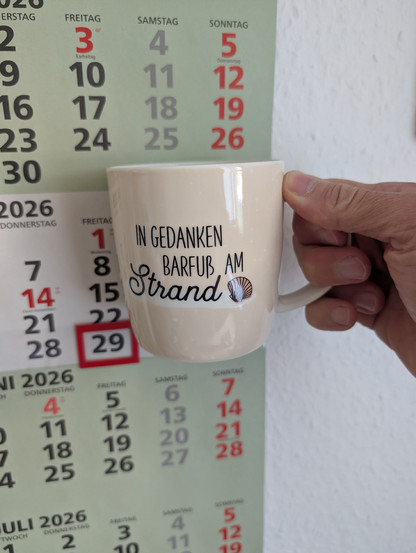 Meine rechte Hand hält einen Porzellan-Kaffeebecher. Seine Farbe ist beige. In schwarzer Schrift steht auf ihm: In Gedanken barfuß am Strand, gefolgt von einer Zeichnung einer bräunlichen Meermuschel. Im Hintergrund hängt ein 3-Monats-Wandkalender an der Wand mit dem rot umrandeten heutigen Tag. 