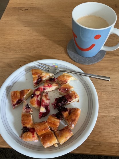 Auf einem hellen Holztisch steht ein weißer Teller mit Teilchen in Stücken. Rechts daneben eine hellblaue Tasse Kaffee mit dem lächelnden, roten Firmenlogo eines Reiseveranstalters.