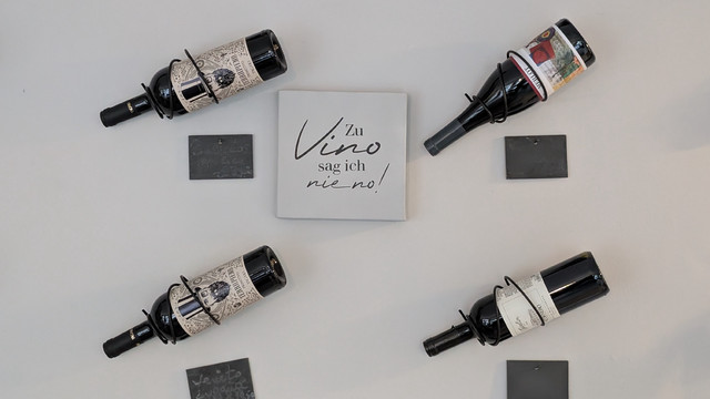 An einer weißen Wand sind Weinflaschen angeschraubt.
Mittig ein Schild mit der Aufschrift "Zu Vino sag ich nie No". 