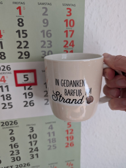 Meine rechte Hand hält einen Porzellan-Kaffeebecher. Seine Farbe ist beige. In schwarzer Schrift steht auf ihm: In Gedanken barfuß am Strand, gefolgt von einer Zeichnung einer bräunlichen Meermuschel. Im Hintergrund hängt ein 3-Monats-Wandkalender an der Wand mit dem rot umrandeten heutigen Tag. 