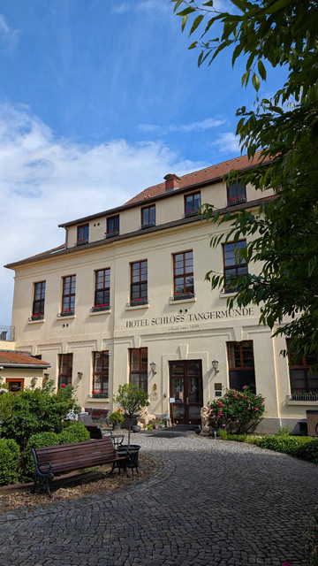 Der halb gepflasterte, halb begrünte Vorhof und das historische Hauptgebäude des Hotels Schloss Tangermünde unter fast blauem Himmel. 
