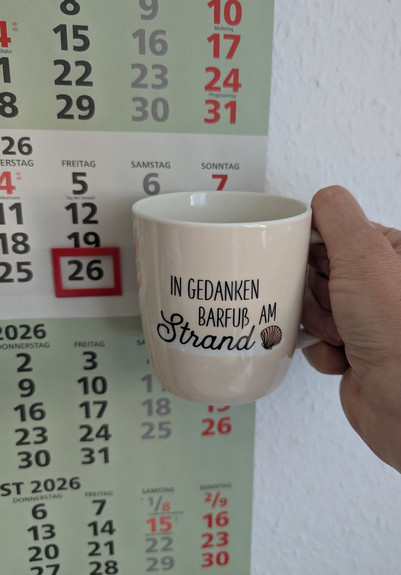 Meine rechte Hand hält einen Porzellan-Kaffeebecher. Seine Farbe ist beige. In schwarzer Schrift steht auf ihm: In Gedanken barfuß am Strand, gefolgt von einer Zeichnung einer bräunlichen Meermuschel. Im Hintergrund hängt ein 3-Monats-Wandkalender an der Wand mit dem rot umrandeten heutigen Tag. 