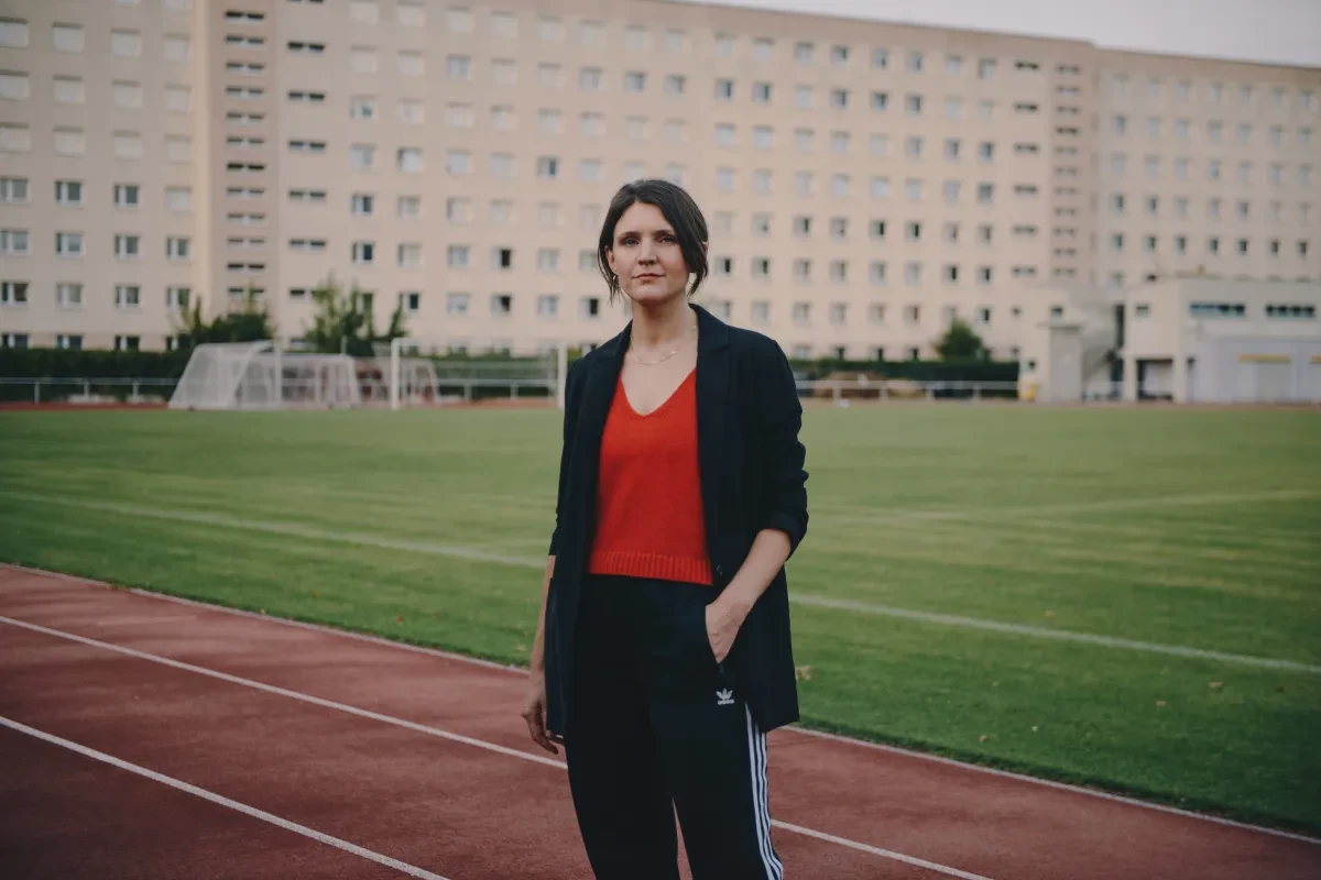 Eine Person mit dunklem, zurückgebundenem Haar steht auf einer Laufbahn vor einem Fußballfeld und blickt in die Kamera. Sie trägt ein rotes Oberteil, ein dunkles Sakko und eine schwarze Trainingshose mit weißen Streifen. Im Hintergrund befindet sich ein großes Mehrfamilienhaus sowie mehrere Fußballtore.