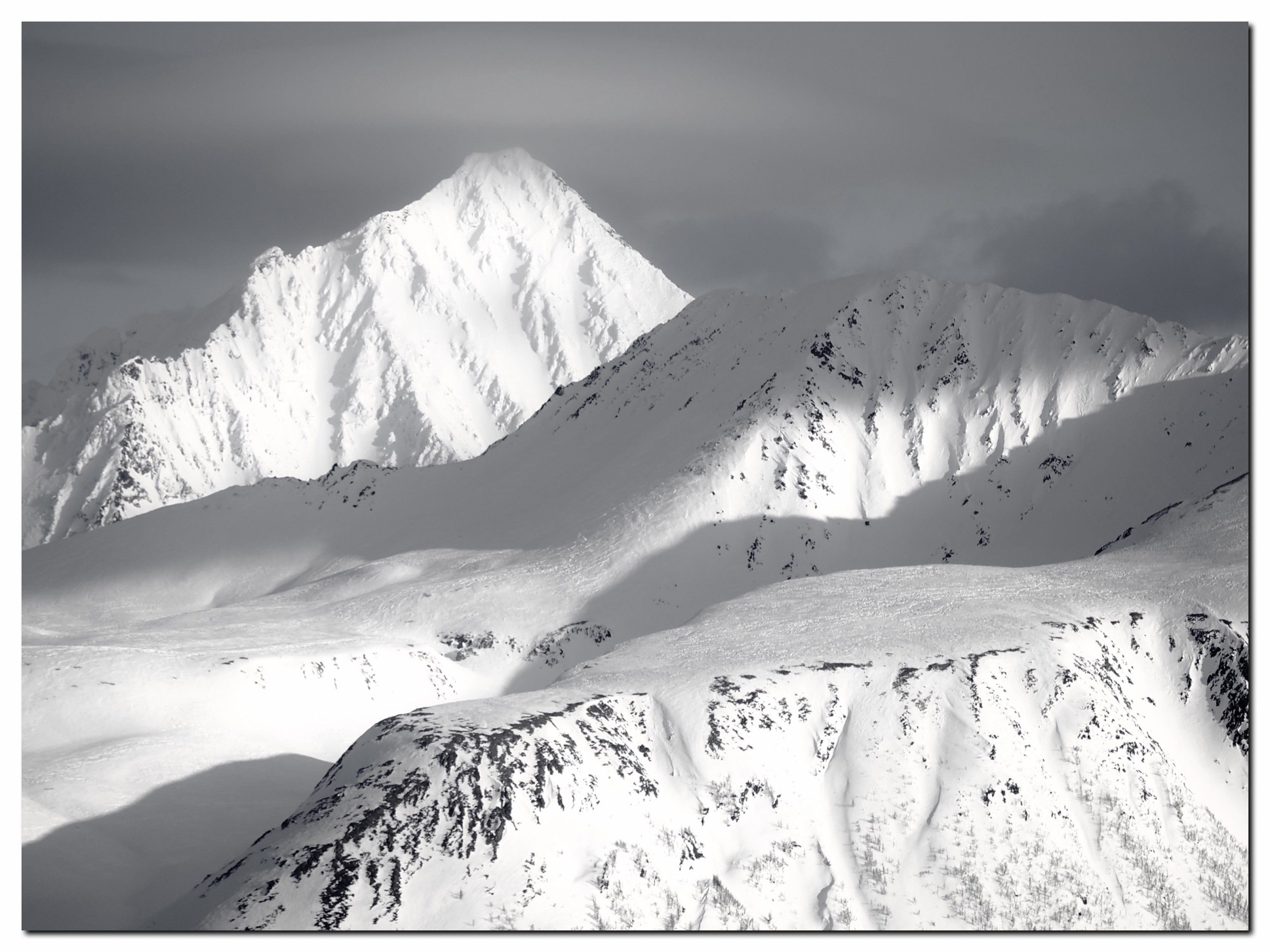 A dramatic black and white landscape featuring snow-covered mountains with peaks and slopes. Shadows create contrast against the bright, sunlit snow. The sky is overcast with a mix of clouds.