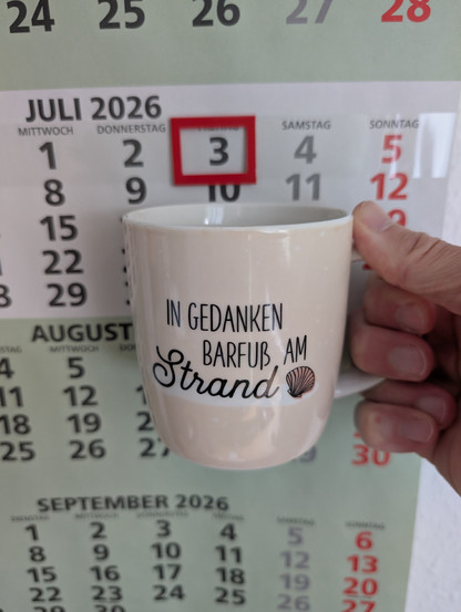Meine rechte Hand hält einen Porzellan-Kaffeebecher. Seine Farbe ist beige. In schwarzer Schrift steht auf ihm: In Gedanken barfuß am Strand, gefolgt von einer Zeichnung einer bräunlichen Meermuschel. Im Hintergrund hängt ein 3-Monats-Wandkalender an der Wand mit dem rot umrandeten heutigen Tag. 