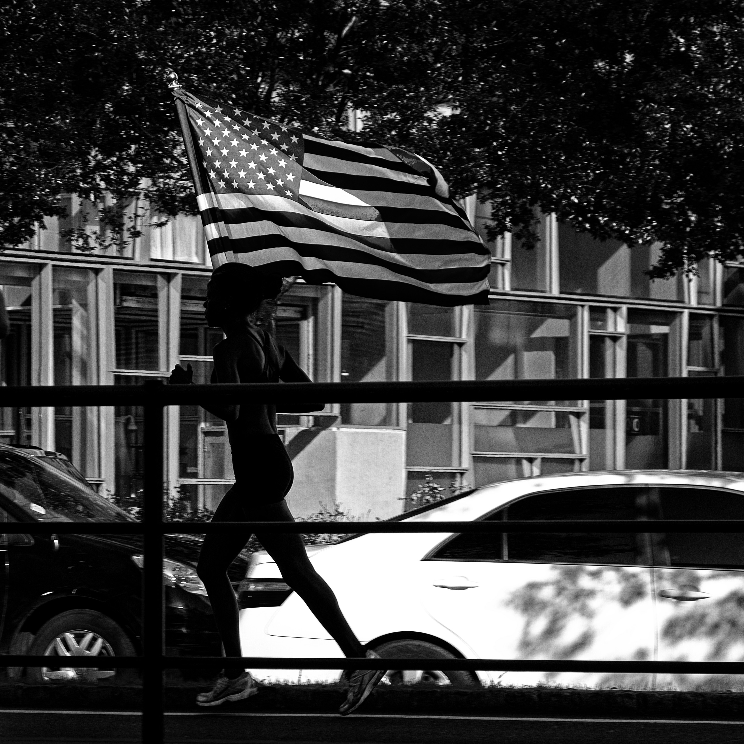 Das quadratische Schwarzweißbild zeigt eine Straßenszene aus New York City. eine junge schwarze Joggerin läuft mit einer wehenden US-Fahne durch das Bild. Während die Joggerin im Schatten liegt, leuchtet die Fahne in der Sonne.