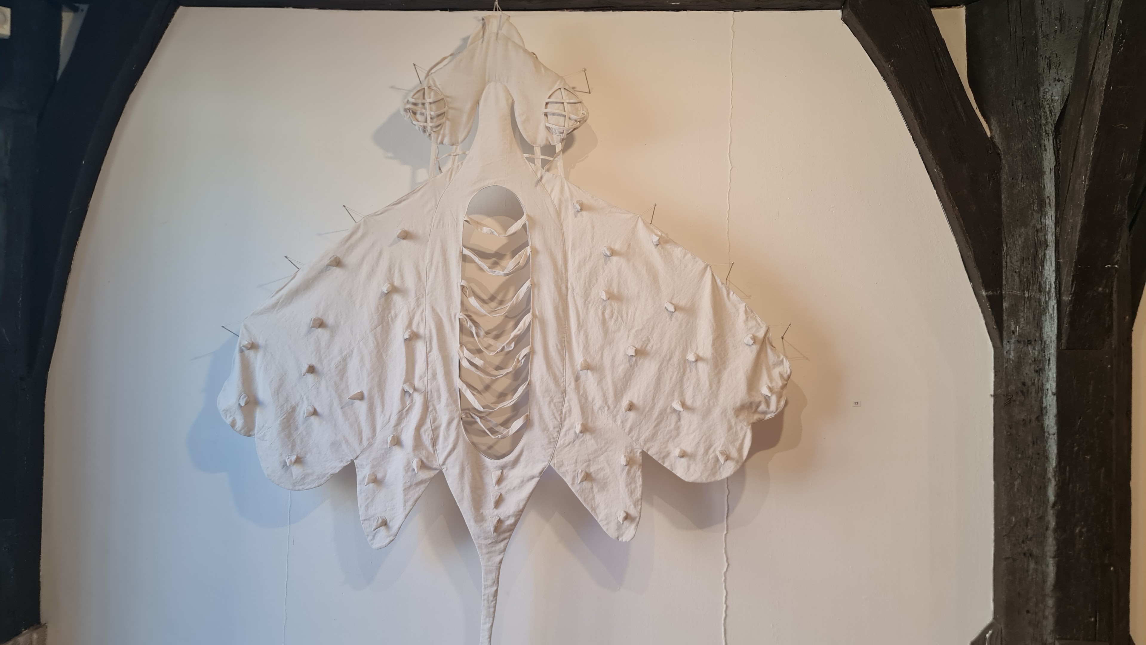 Aus weissem Textilstoff gemachte, genäht und geflochten, insektoide Formen, wie kostüme zum anziehen, hängen als Kunstwerk an weisser Wand, hier ein grosses, nächstes Bild mehrere kleinere nebeneinander