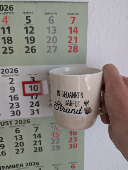 Meine rechte Hand hält einen Porzellan-Kaffeebecher. Seine Farbe ist beige. In schwarzer Schrift steht auf ihm: In Gedanken barfuß am Strand, gefolgt von einer Zeichnung einer bräunlichen Meermuschel. Im Hintergrund hängt ein 3-Monats-Wandkalender an der Wand mit dem rot umrandeten heutigen Tag. 