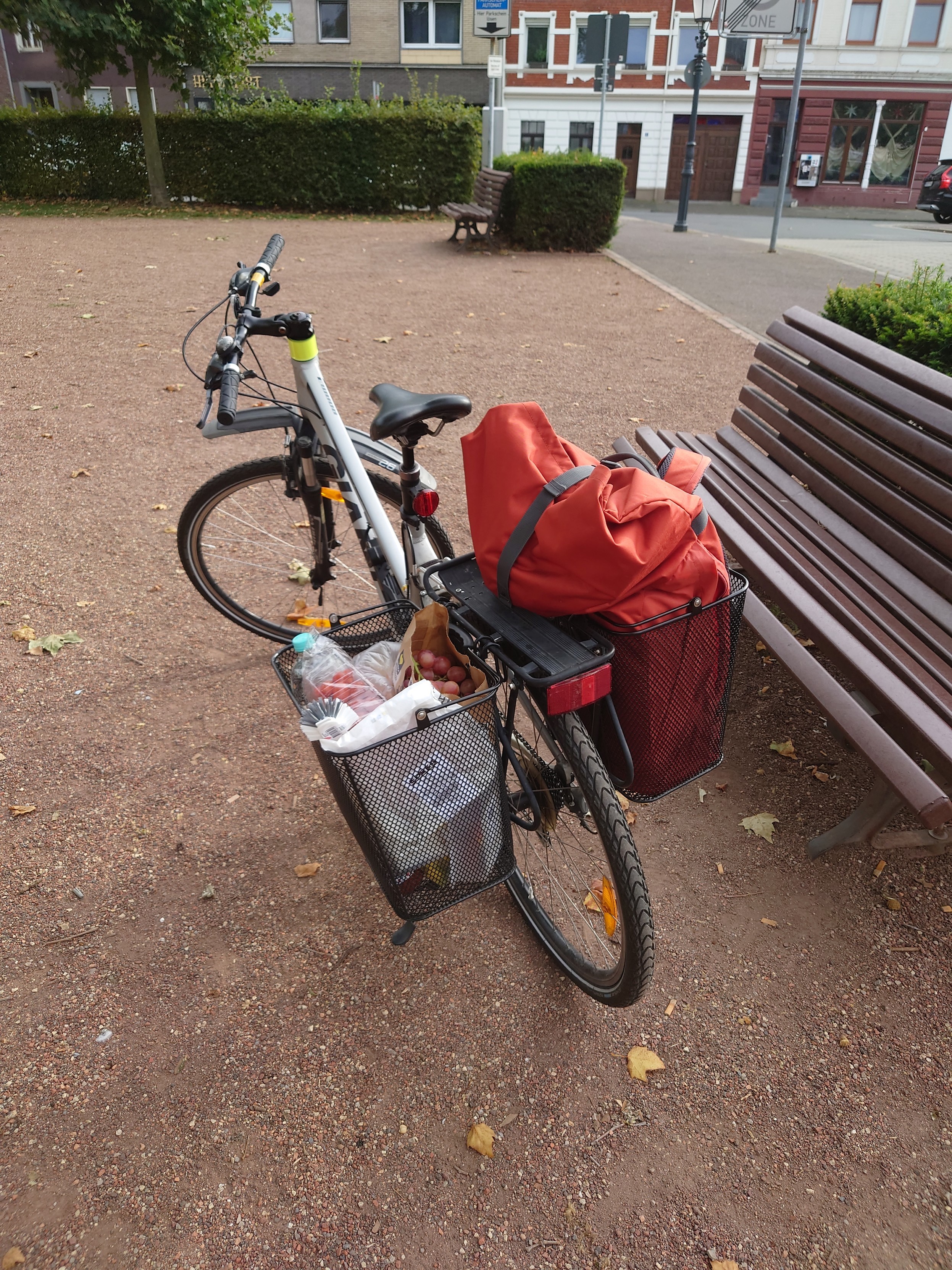 a bike with two metal baskets on the rear rack. One carries some groceries and there is a large red bag in the other.