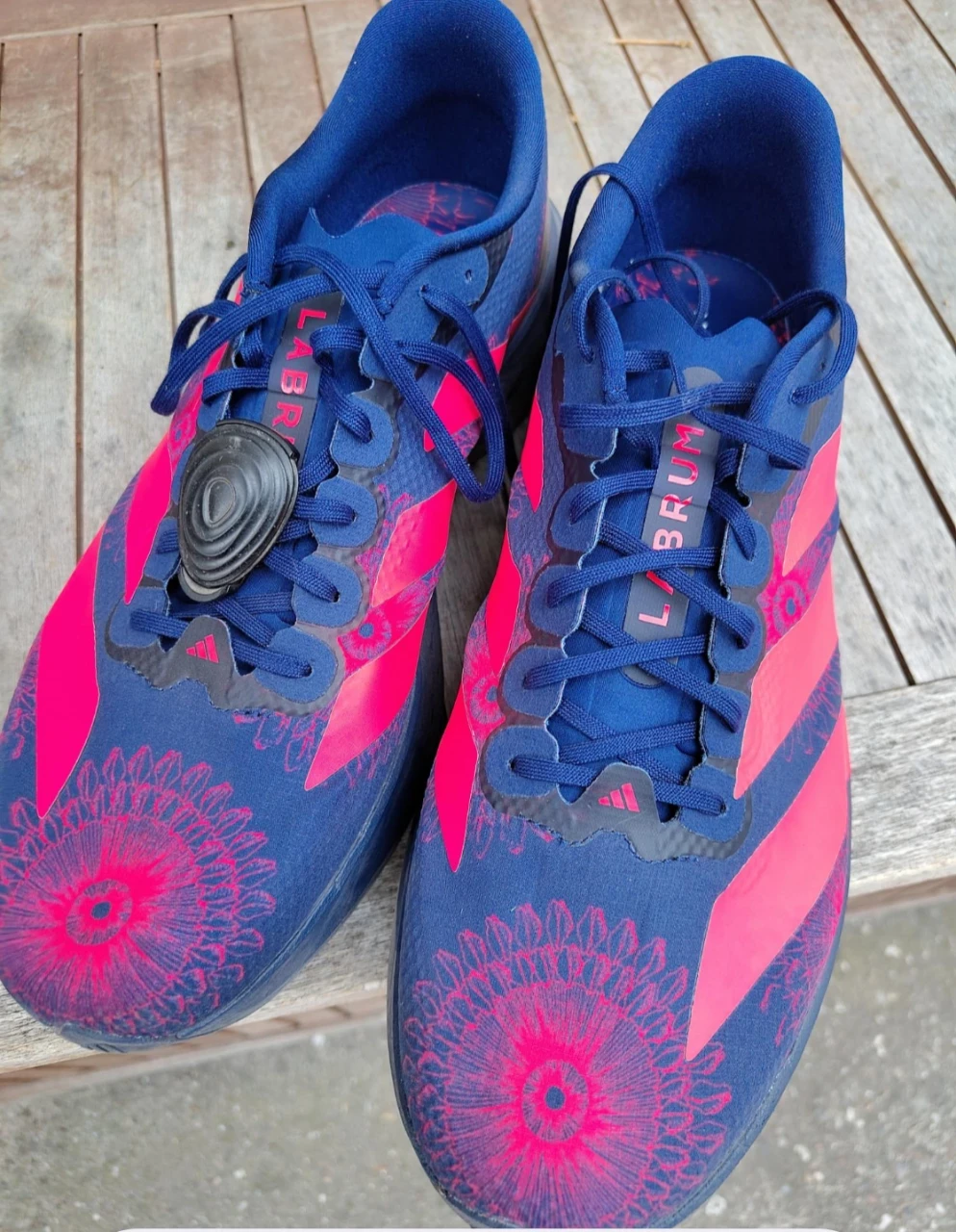 A pair of blue and pink Adidas running shoes with a floral pattern, placed on a wooden surface. The shoes have the word "LABRUM" printed on the tongue and feature a unique lacing system with a black circular lace lock. The Adidas logo is visible on the side.