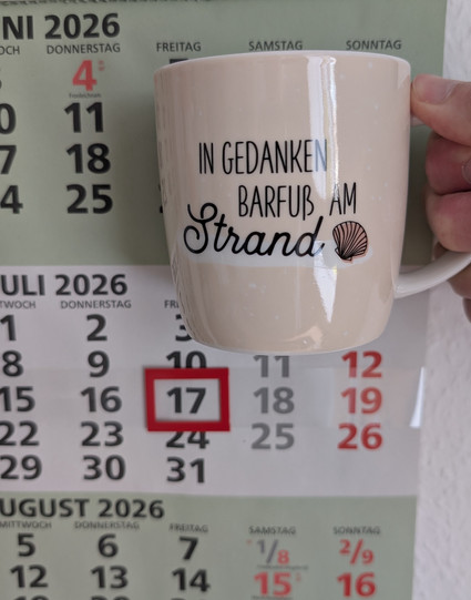 Meine rechte Hand hält einen Porzellan-Kaffeebecher. Seine Farbe ist beige. In schwarzer Schrift steht auf ihm: In Gedanken barfuß am Strand, gefolgt von einer Zeichnung einer bräunlichen Meermuschel. Im Hintergrund hängt ein 3-Monats-Wandkalender an der Wand mit dem rot umrandeten heutigen Tag. 