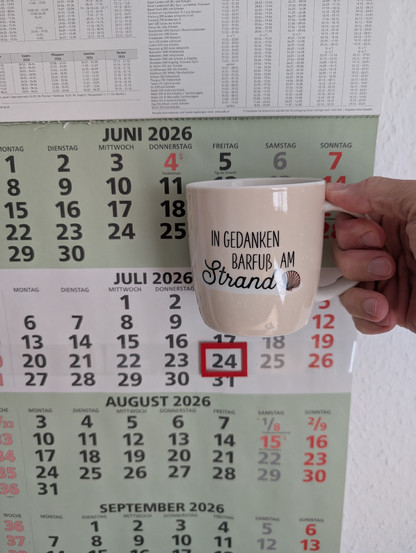 Meine rechte Hand hält einen Porzellan-Kaffeebecher. Seine Farbe ist beige. In schwarzer Schrift steht auf ihm: In Gedanken barfuß am Strand, gefolgt von einer Zeichnung einer bräunlichen Meermuschel. Im Hintergrund hängt ein 3-Monats-Wandkalender an der Wand mit dem rot umrandeten heutigen Tag. 