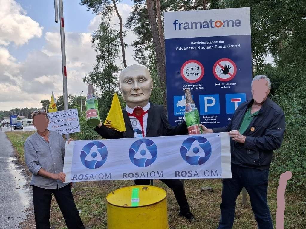 Zwei Demonstranten mit einem Transparent mit Rosatom-Logo vor der Brennelementefabrik Lingen mit einem verkleideten Putin, der vor dem Betreiber-Firmenschild von framatome symbolisch die Sektkorken knallen lässt. Im Vordergrund ein symbolisches Atommüllfass