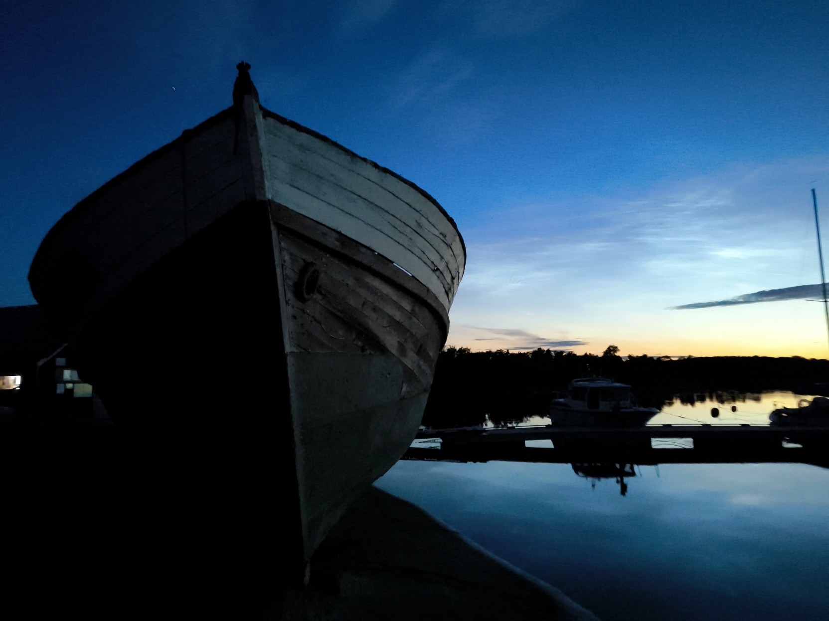 A large, dark wooden boat dominates the left foreground of this twilight scene. The bow of the vessel faces toward the viewer, showing textured planks in shadow. The sky transitions from deep blue at the top to a pale yellow near the horizon line. In the distance, a dark treeline sits along the edge of the water, with the silhouettes of other small boats and a pier visible. Reflections of the sky and structures appear on the calm surface of the water.