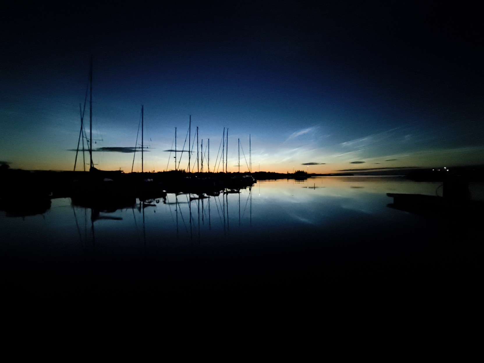 A silhouette of several sailboats docked in a calm body of water during twilight. The sky transitions from deep blue at the top to light yellow and orange near the horizon. Multiple sailboat masts and hulls appear as dark shapes against the lighter part of the sky. The still surface of the water reflects the vertical lines of the masts and the colors from the horizon. Wispy clouds are scattered across the middle section of the sky.