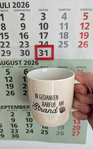 Meine rechte Hand hält einen Porzellan-Kaffeebecher. Seine Farbe ist beige. In schwarzer Schrift steht auf ihm: In Gedanken barfuß am Strand, gefolgt von einer Zeichnung einer bräunlichen Meermuschel. Im Hintergrund hängt ein 3-Monats-Wandkalender an der Wand mit dem rot umrandeten heutigen Tag. 