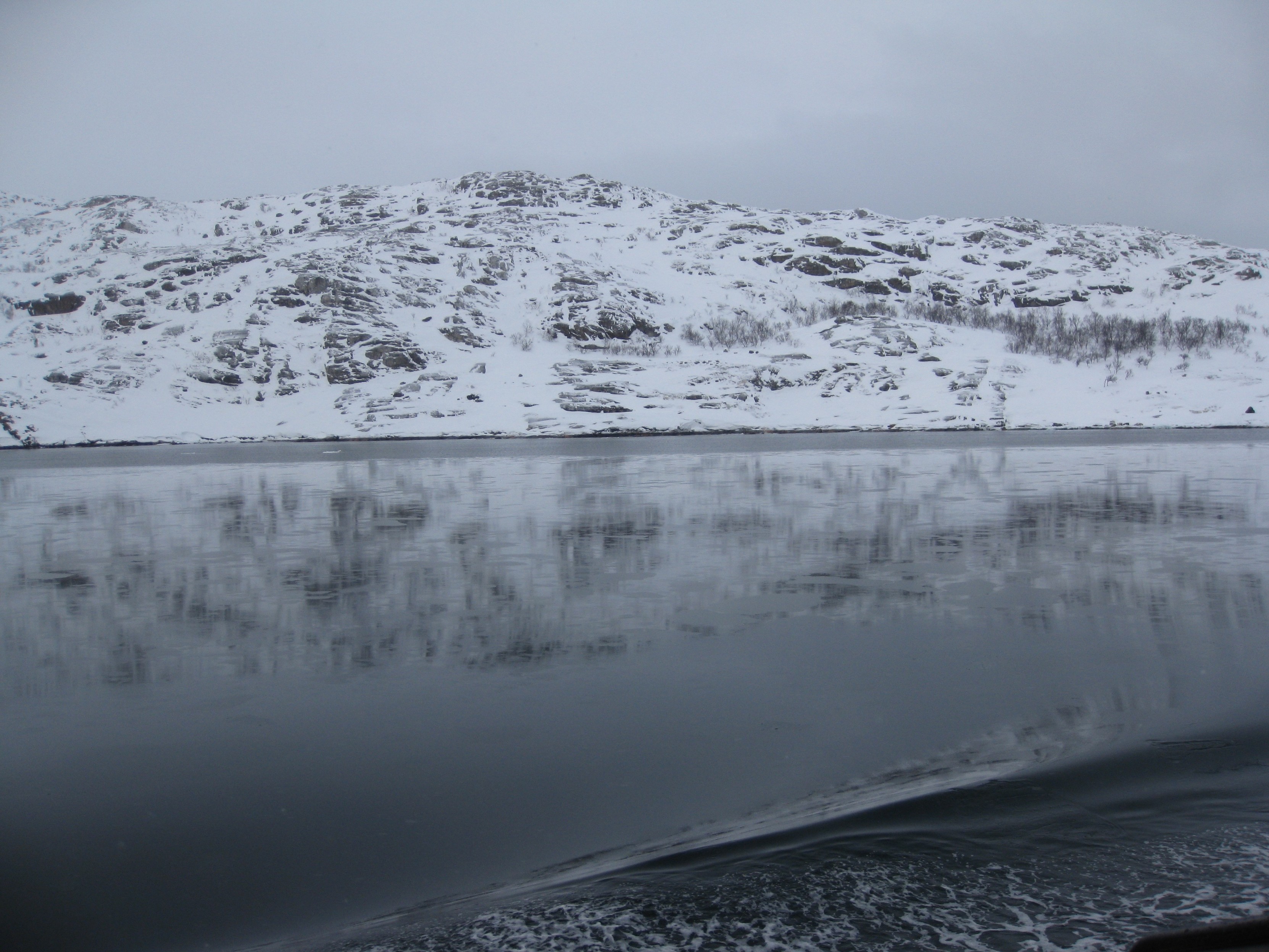 Eine schneebedeckte Berglandschaft, die sich in ruhigem Wasser spiegelt, mit bewölktem Himmel im Hintergrund.
