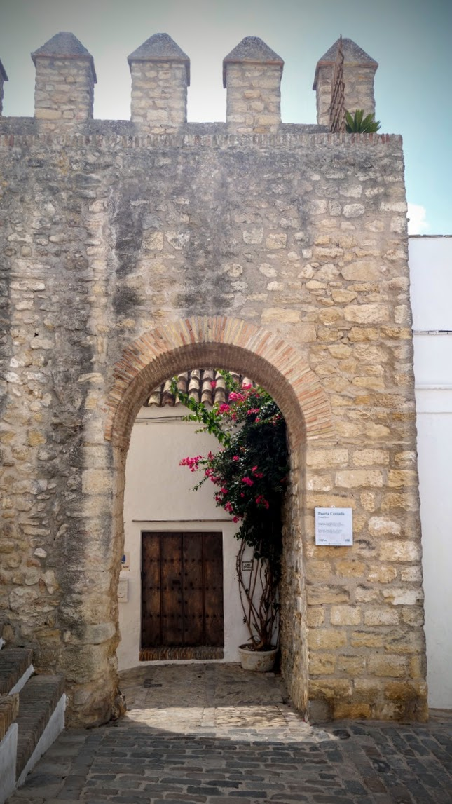 Historisches steinernes Tor mit Zinnen und rundem Backsteinbogen. Durch den Torbogen sieht man eine weiß verputzte Hausfassade mit einer dunklen Holztür. Rechts daneben wächst ein üppiger Bougainvillea-Strauch mit leuchtend pinken Blüten. Der Boden ist mit Kopfsteinpflaster ausgelegt, die Szene wirkt ruhig und mediterran.