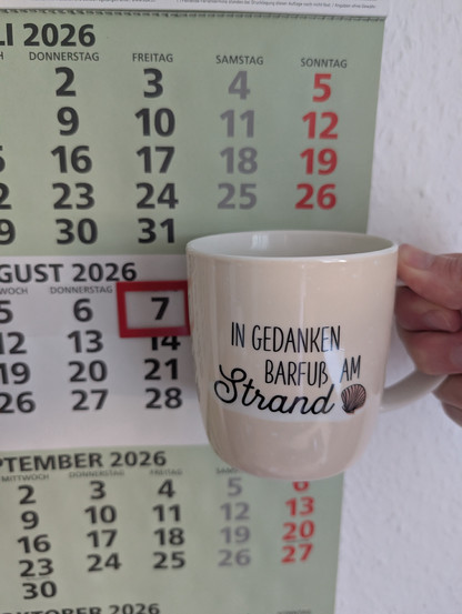 Meine rechte Hand hält einen Porzellan-Kaffeebecher. Seine Farbe ist beige. In schwarzer Schrift steht auf ihm: In Gedanken barfuß am Strand, gefolgt von einer Zeichnung einer bräunlichen Meermuschel. Im Hintergrund hängt ein 3-Monats-Wandkalender an der Wand mit dem rot umrandeten heutigen Tag. 
