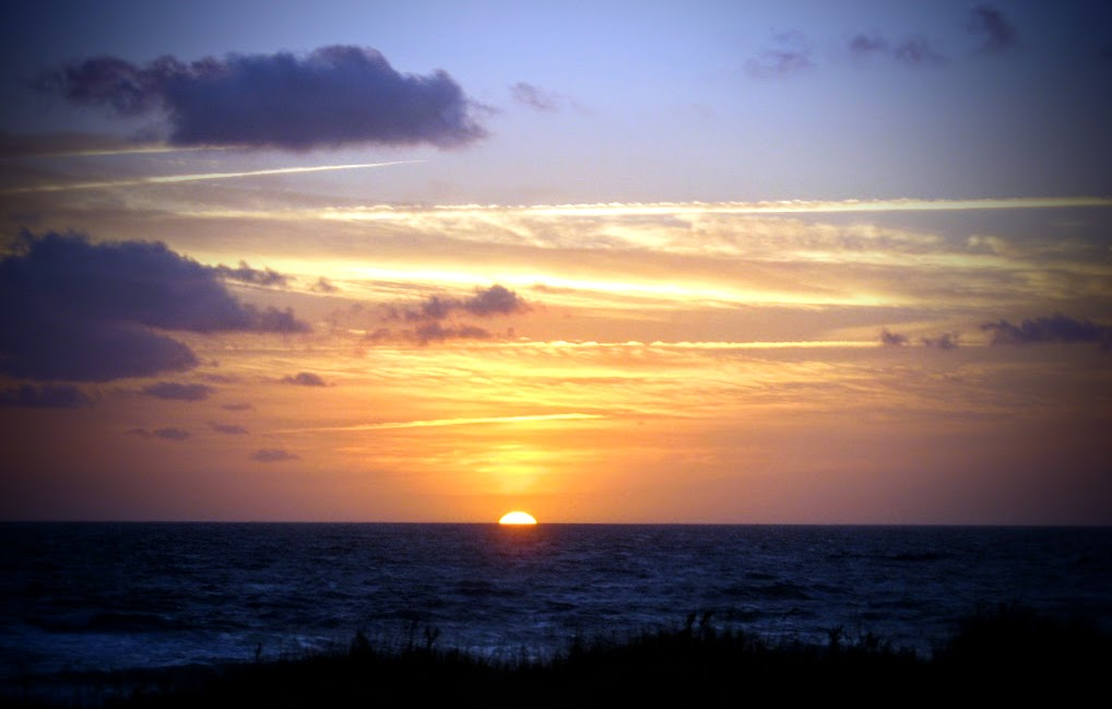 Am Horizont ist die Sonne gerade dabei, im Meer zu versinken. Nur ein kleiner Bogen ist noch sichtbar. Der Himmel leuchtet in warmen Gelb-, Orange- und Rosatönen, darüber ziehen sich dunkle Wolken. Im Vordergrund zeichnet sich die Küste als dunkle Silhouette ab, während das Meer ruhig und dunkelblau bis zum Horizont reicht.