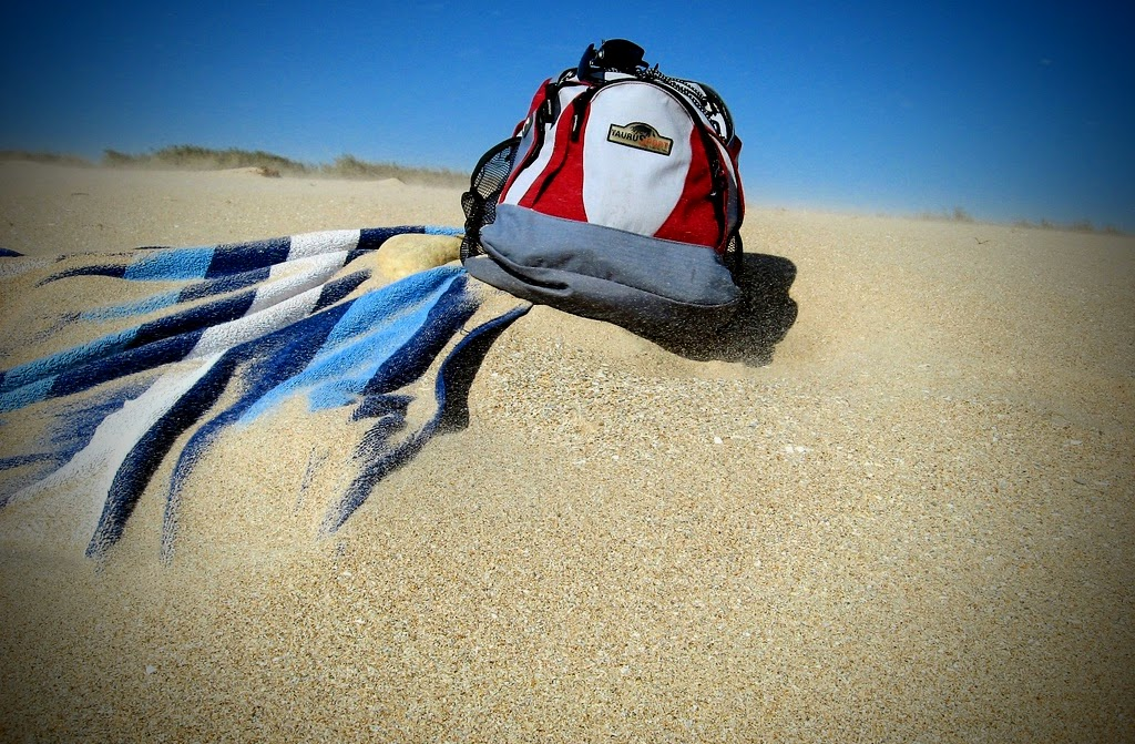 Ein Rucksack liegt auf einem weitläufigen, sonnigen Sandstrand. Neben ihm liegt ein blau-weiß gestreiftes Strandtuch, das teilweise im Sand verweht ist. Feine Sandkörner werden vom Wind aufgewirbelt und ziehen als zarte, sandige Staubfahne über den Boden, besonders hinter und links vom Rucksack. Im Hintergrund sind niedrige Dünen zu erkennen, darüber erstreckt sich ein klarer, tiefblauer Himmel.
