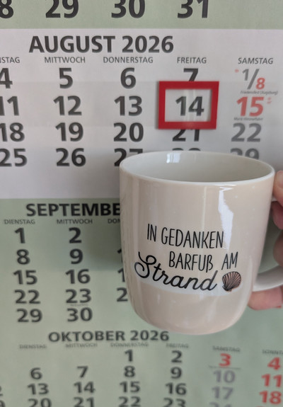 Meine rechte Hand hält einen Porzellan-Kaffeebecher. Seine Farbe ist beige. In schwarzer Schrift steht auf ihm: In Gedanken barfuß am Strand, gefolgt von einer Zeichnung einer bräunlichen Meermuschel. Im Hintergrund hängt ein 3-Monats-Wandkalender an der Wand mit dem rot umrandeten heutigen Tag. 