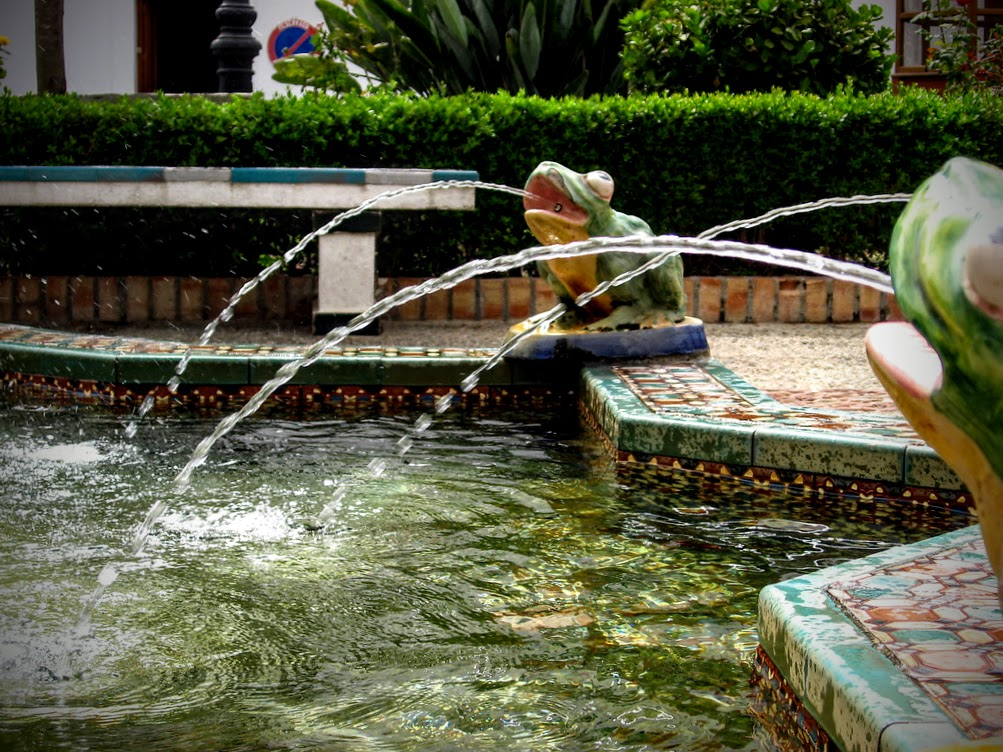 Ein dekorativer Brunnen mit mehreren Froschfiguren aus Keramik. Aus den Mäulern der Frösche spritzen Wasserstrahlen in ein großes, mit bunten Fliesen eingefasstes Becken. Im Hintergrund sind eine gepflegte Hecke und Pflanzen zu sehen.