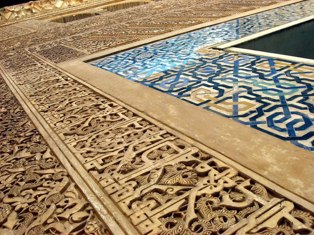 Detail der Nasridenpaläste der Alhambra: Aufwendig geschnitzte Stein- und Stuckornamente mit dicht ineinander verschlungenen floralen und geometrischen Mustern. Daneben ist ein blau-weißes Mosaik aus geometrisch verschlungenen Formen zu sehen. Die Aufnahme zeigt die kunstvoll verzierten Flächen aus einer schrägen Nahperspektive.