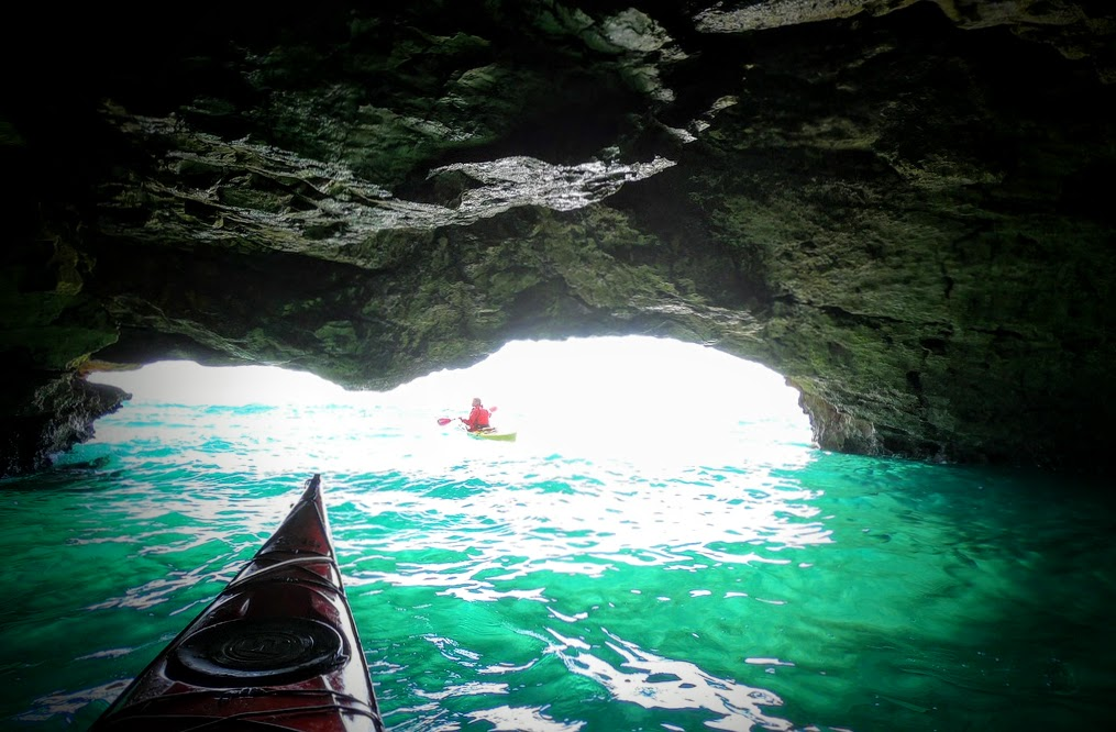 Blick aus einem Kajak in einer felsigen Höhle am Meer. Das dunkle Felsdach spendet Schatten und umrahmt den hellen Höhlenausgang. Innen leuchtet smaragdgrünes, leicht bewegtes Wasser. In der Ferne paddelt eine weitere Person in einem Kajak