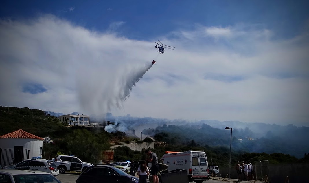 Über einem von Rauch und dichtem Qualm bedeckten, bewaldeten Hang fliegt ein Löschhubschrauber und wirft einen Wasserstrahl auf den Brandherd. Im Vordergrund stehen mehrere Einsatzfahrzeuge, während Menschen die Löscharbeiten beobachten. Im Hintergrund sind weitere Rauchschwaden und Gebäude am Hang zu sehen. Mittendrin auch Feuerwehrfahrzeuge auf einem Forstweg.