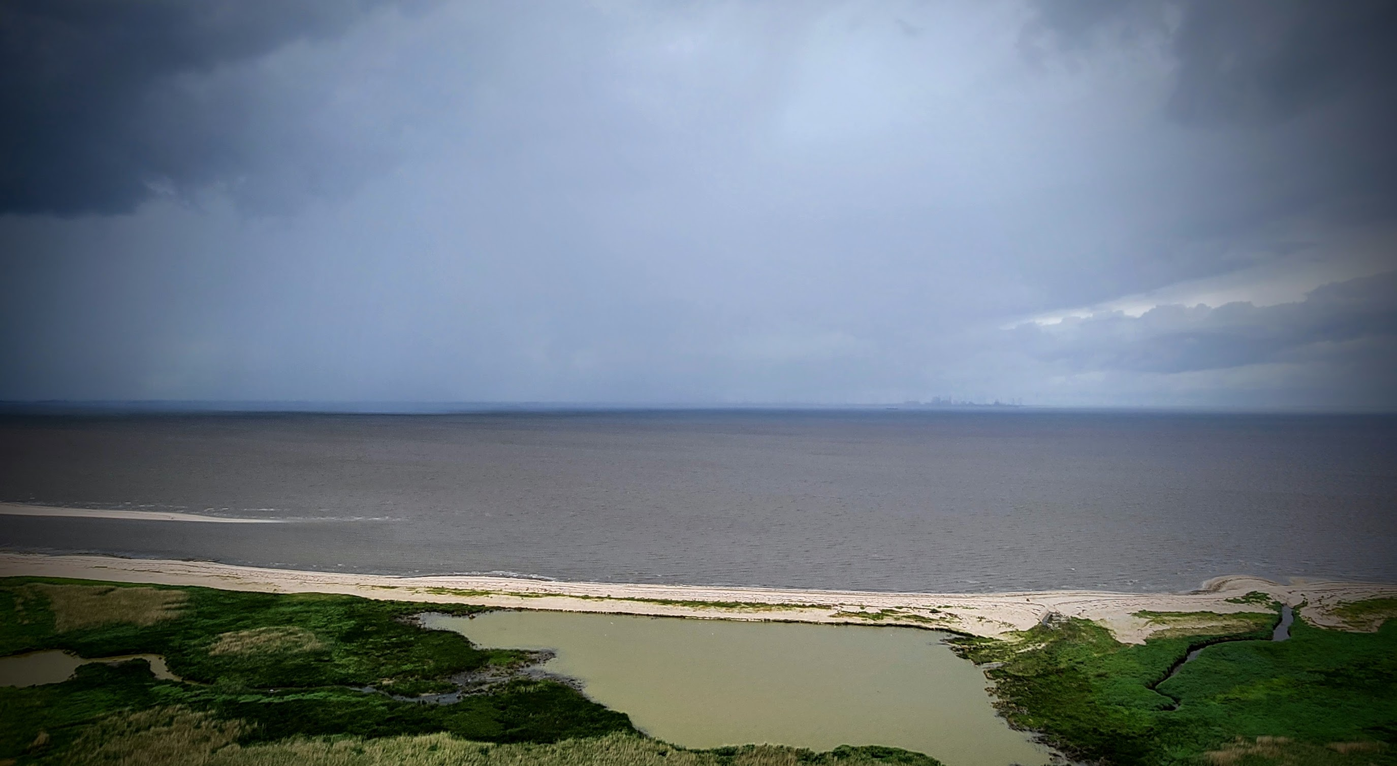 Blick über eine flache Küstenlandschaft auf das ruhige Meer. Im Vordergrund liegen grüne Salzwiesen und ein flaches, grünlich-braunes Gewässer, dahinter ein schmaler heller Sandstrand. Am Horizont ist unter einem dunklen, wolkenverhangenen Himmel schemenhaft eine Stadt zu erkennen. Die Wolkendecke reißt rechts etwas auf und lässt helles Licht auf das Meer fallen.