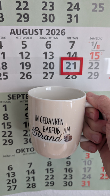 Meine rechte Hand hält einen Porzellan-Kaffeebecher. Seine Farbe ist beige. In schwarzer Schrift steht auf ihm: In Gedanken barfuß am Strand, gefolgt von einer Zeichnung einer bräunlichen Meermuschel. Im Hintergrund hängt ein 3-Monats-Wandkalender an der Wand mit dem rot umrandeten heutigen Tag. 