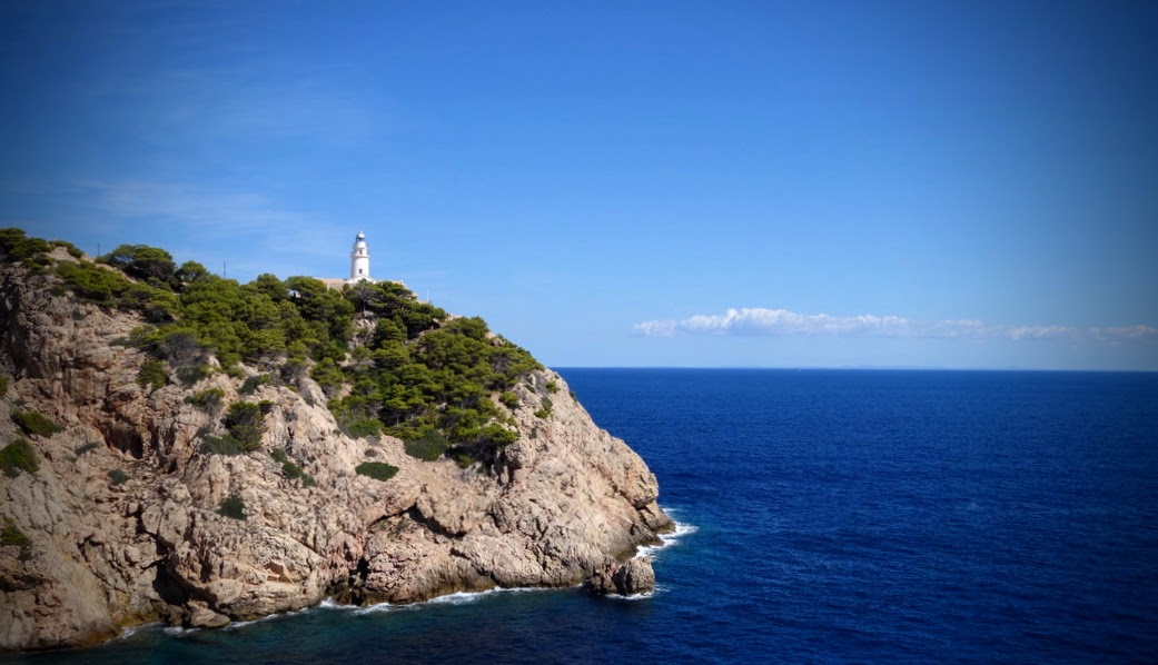 Eine felsige Küste mit dicht bewachsenen grünen Bäumen und Sträuchern ragt ins tiefblaue Meer. Auf der Anhöhe steht ein kleiner weißer Leuchtturm. Über dem ruhigen Horizont erstreckt sich ein klarer, wolkenarmer blauer Himmel.