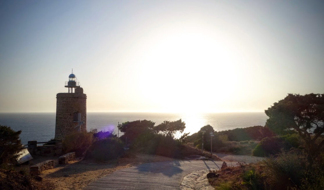 Ein kleiner, runder Leuchtturm aus dunklem Naturstein steht links auf einer bewachsenen Küstenhöhe. Die Sonne steht tief über dem Atlantik und spiegelt sich auf dem Wasser. Ein schmaler Weg führt durch die karge, von niedrigen Bäumen und Sträuchern bewachsene Landschaft zum Leuchtturm. Die tief stehende Sonne erzeugt starke Gegenlichtstimmung und lange Schatten.