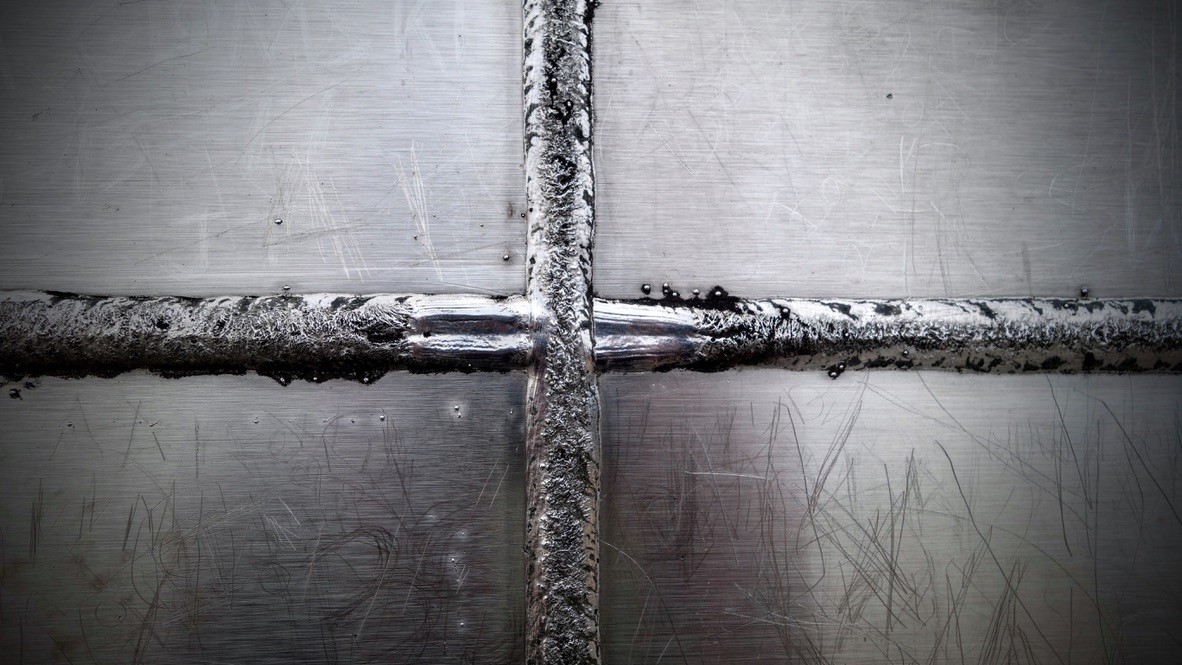 Nahaufnahme einer verschweißten Metalloberfläche. Zwei breite, silbrig-graue Schweißnähte kreuzen sich in der Bildmitte. Die Metallfläche ist stark zerkratzt und weist dunkle sowie helle Verfärbungen und kleine Schweißspritzer auf. Das Bild ist überwiegend in Grau- und Schwarztönen gehalten.