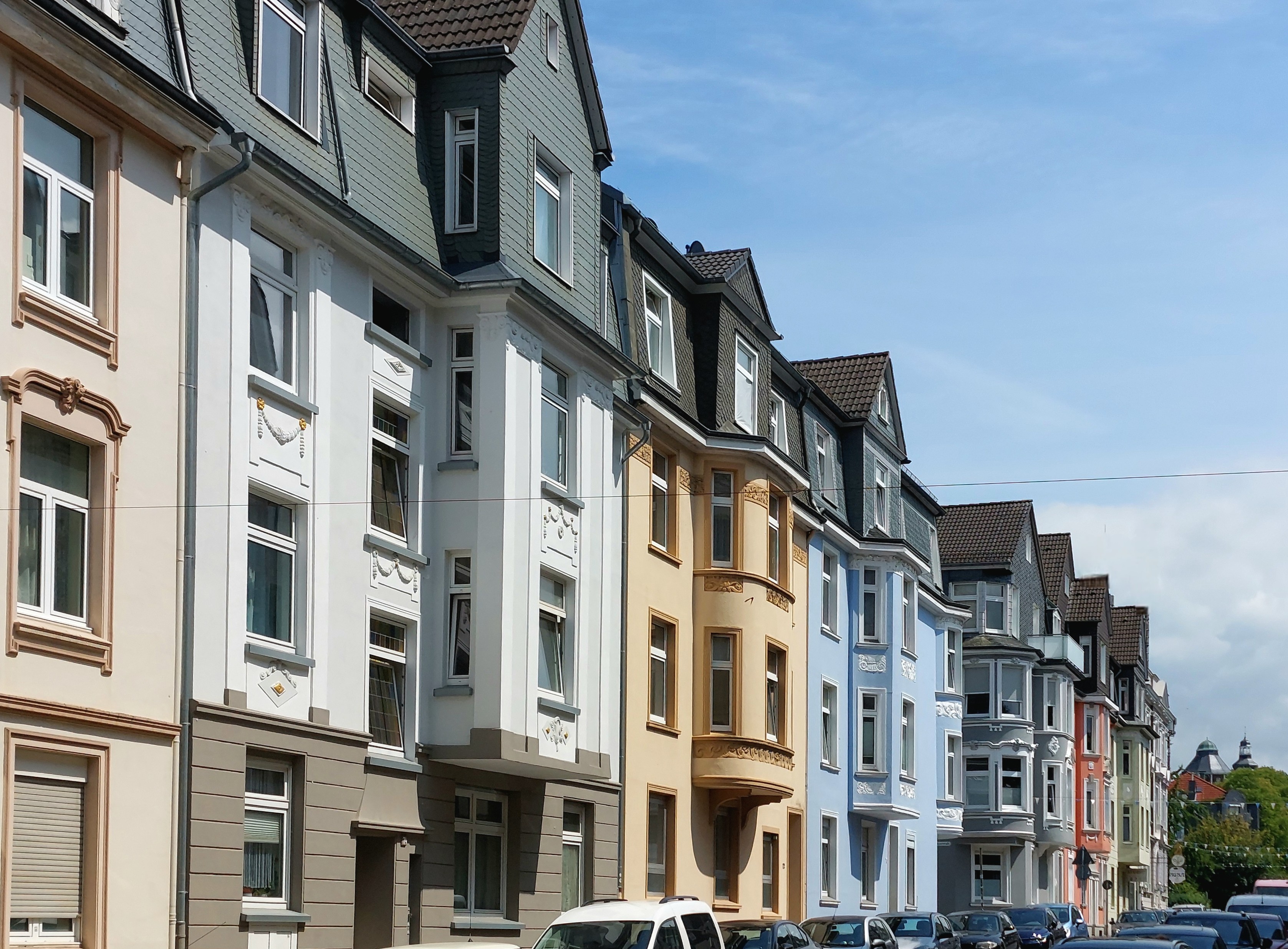 Bunte Häuserzeile der Hindenburgstraße in der Sonne, mit unterschiedlich geformten Erkern. Fenster ohne Ende. Blauer Himmel mit Wolken. Von den vielen geparkten Fahrzeugen sieht man fast nur die Dächer.
