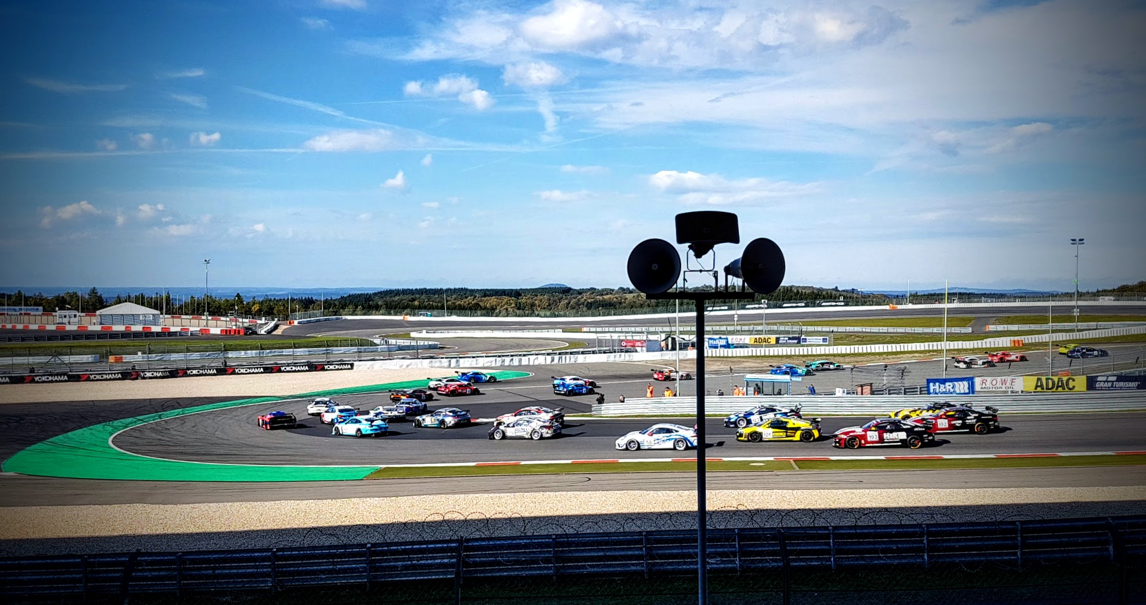 Rennstrecke bei sonnigem Wetter: Eine große Gruppe von Tourenwagen fährt dicht beieinander durch eine Kurve. Die Fahrzeuge sind unterschiedlich lackiert, unter anderem gelb, rot, weiß, blau und türkis. Im Vordergrund stehen ein hoher Lautsprecher- oder Kameramast und eine Leitplanke. Im Hintergrund sind weitere Streckenabschnitte, Tribünen und Gebäude sowie eine bewaldete Landschaft unter blauem Himmel zu sehen.