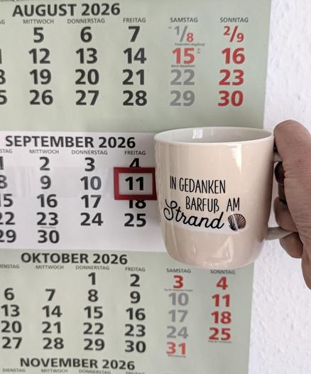 Meine rechte Hand hält einen Porzellan-Kaffeebecher. Seine Farbe ist beige. In schwarzer Schrift steht auf ihm: In Gedanken barfuß am Strand, gefolgt von einer Zeichnung einer bräunlichen Meermuschel. Im Hintergrund hängt ein 3-Monats-Wandkalender an der Wand mit dem rot umrandeten heutigen Tag.