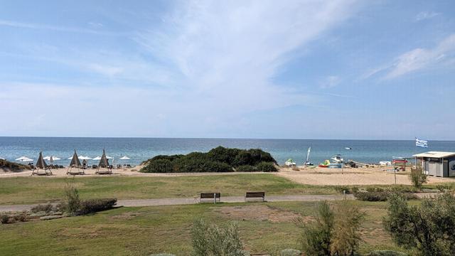 Das Foto zeigt einen sonnigen Ausblick auf einen weitläufigen Sandstrand und das ruhige, blau schimmernde Meer unter einem leicht bewölkten Himmel. Im Vordergrund erstreckt sich eine gepflegte grüne Rasenfläche mit vereinzelten Büschen, einem Fußweg und einer Holzbank. Am hellen Strand dahinter sind mehrere weiße Sonnenschirme sowie drei markante, pyramidenförmige Strohschirme mit Liegen aufgestellt, während der rechte Strandabschnitt von grünen Dünenbüschen begrenzt wird. Die erhöhte Perspekti…