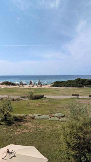 Das Foto zeigt einen sonnigen Ausblick auf einen weitläufigen Sandstrand und das ruhige, blau schimmernde Meer unter einem leicht bewölkten Himmel. Im Vordergrund erstreckt sich eine gepflegte grüne Rasenfläche mit vereinzelten Büschen, einem Fußweg und einer Holzbank. Am hellen Strand dahinter sind mehrere weiße Sonnenschirme sowie drei markante, pyramidenförmige Strohschirme mit Liegen aufgestellt, während der rechte Strandabschnitt von grünen Dünenbüschen begrenzt wird. Die erhöhte Perspekti…